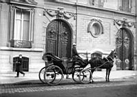 Femme cocher et son taxi hippomobile devant deux portes cocheres de la fin du XVIIIeme siecle. Paris, vers 1900